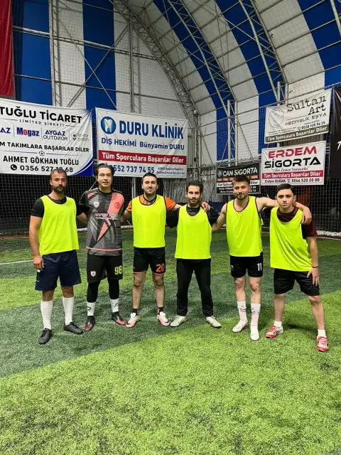 Mavi-beyaz çatılı kapalı halı sahada, tribün pankartlarının önünde takım fotoğrafı.